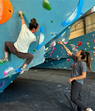 Two people at an Austin Climbing Gym: one climbs a blue route on an overhanging wall, while the other stands below, pointing up and offering guidance. Brightly colored climbing holds are visible on the wall.