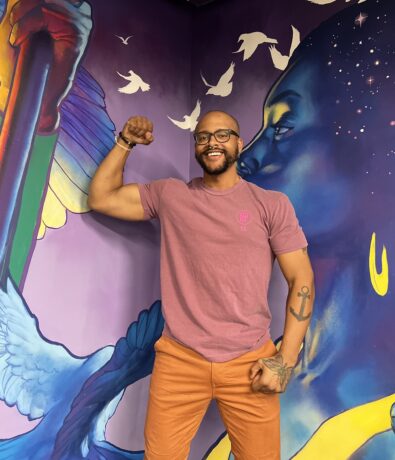 A smiling person with glasses poses by flexing their arm in front of a colorful mural featuring a blue bird, white doves, and a large profile of a person with deep blue skin and yellow earrings, evoking an uplifting, climbing spirit.