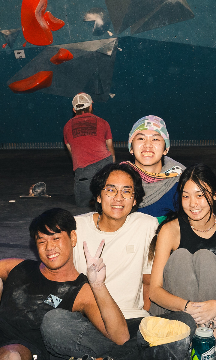 Four young people sit on the floor of a Salt Lake City climbing gym, smiling and posing for the camera. One flashes a peace sign. Climbing holds and another member facing the wall are visible in the background.