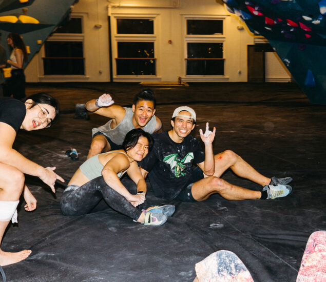 Four young adults with climbing gear and chalk on their hands pose and smile while sitting on the floor of an indoor climbing gym, enjoying Membership Benefits at Austin Bouldering Project, surrounded by colorful climbing walls and holds.