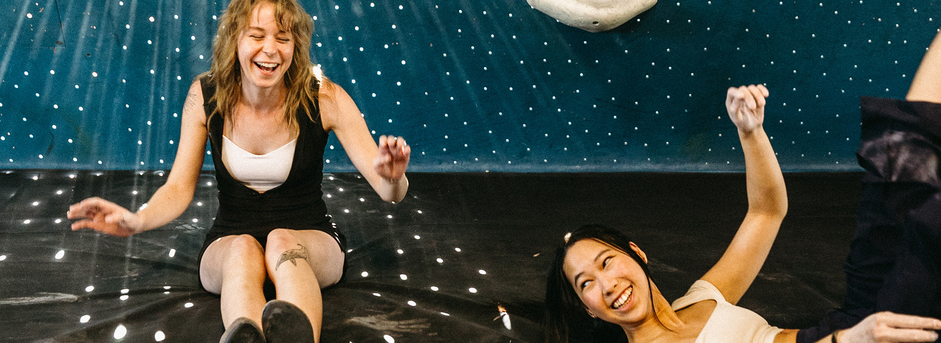 Two women sit and laugh together on a padded gym floor in Salt Lake City, appearing joyful and relaxed, with a blue climbing wall featuring small white holds and dotted patterns behind them. Enjoy 24/7 access to this vibrant space.