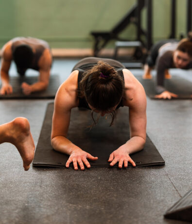 People exercising indoors on yoga mats; the focus is on a woman in a plank position with her forearms and toes on the mat, others in similar positions in the background.