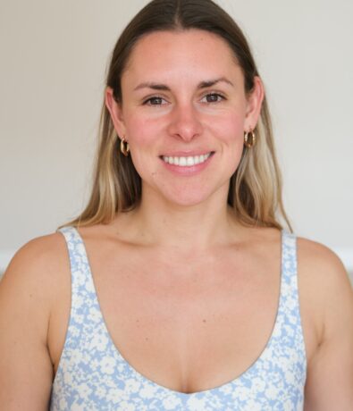 A woman with long, light brown hair wearing gold hoop earrings and a blue and white floral top smiles at the camera against a plain light background, embodying the vibrant energy of Brooklyn Climbing Yoga and Fitness.