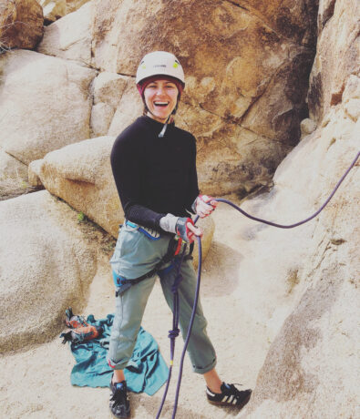 A smiling person wearing a helmet, gloves, and climbing gear stands on sandy ground near large rocks at Upper Walls in Fremont, holding a rope and looking up.