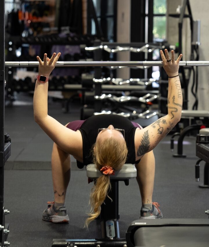 A woman with a ponytail and tattoos lies on a bench, preparing to lift a barbell in a gym filled with workout and climbing equipment. Another person runs past in the foreground, adding to the energetic scene.