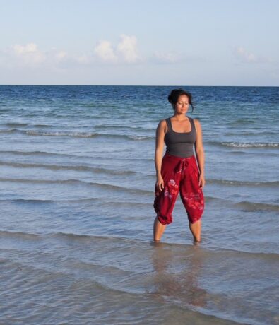 A woman in a gray tank top and red pants stands ankle-deep in the shallow water at Fremont Beach, looking calm under a clear sky with gentle waves and the upper walls of nearby cliffs in the background.