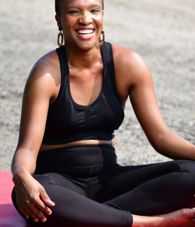 A smiling woman in a black sports outfit sits cross-legged on a red exercise mat outdoors.