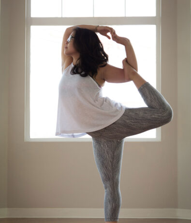 A woman practices yoga indoors near the upper walls of her Fremont home, balancing on one leg in the dancer pose by a large window, wearing a white tank top and patterned leggings.