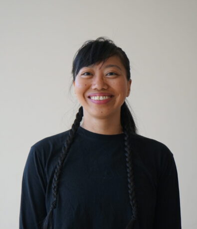 Headshot of Nikki, with long black braids, wearing a black long-sleeve crop top and leopard-print leggings, smiles at the camera against a plain light background, capturing the vibrant spirit of the Yoga Room.