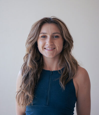 headshot of Emily O. She has long brown hair and is wearing a sleeveless dark blue top. In front of a plain light-colored background, smiling at the camera.