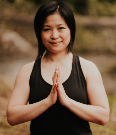 A woman with short black hair sits outdoors, wearing a black tank top. She is holding her hands together in a prayer position and smiling gently, with greenery in the blurred background.
