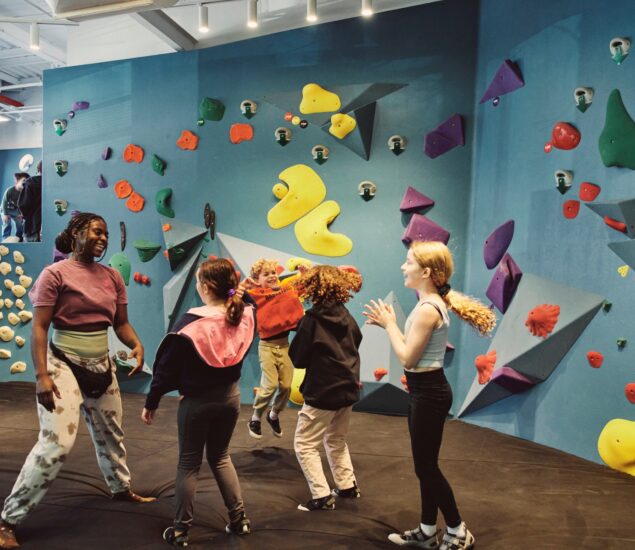 A group of children and an adult stand and talk near colorful climbing walls in a Brooklyn indoor bouldering gym. Wearing climbing shoes and casual athletic clothing, theyre ready for 2024 Summer Adventures among the bright holds.