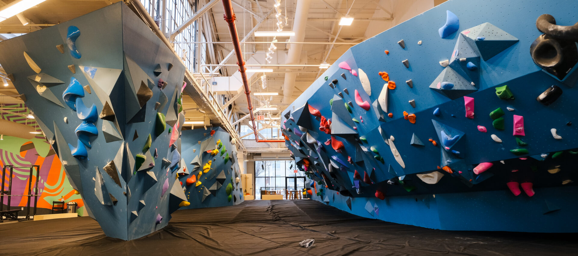 Brooklyn Climbing Yoga and Fitness offers an indoor rock climbing gym with large blue walls, colorful holds, padded black flooring, and bright lighting. Exposed ceiling beams and windows on one wall create an open, inviting space. No people are present.