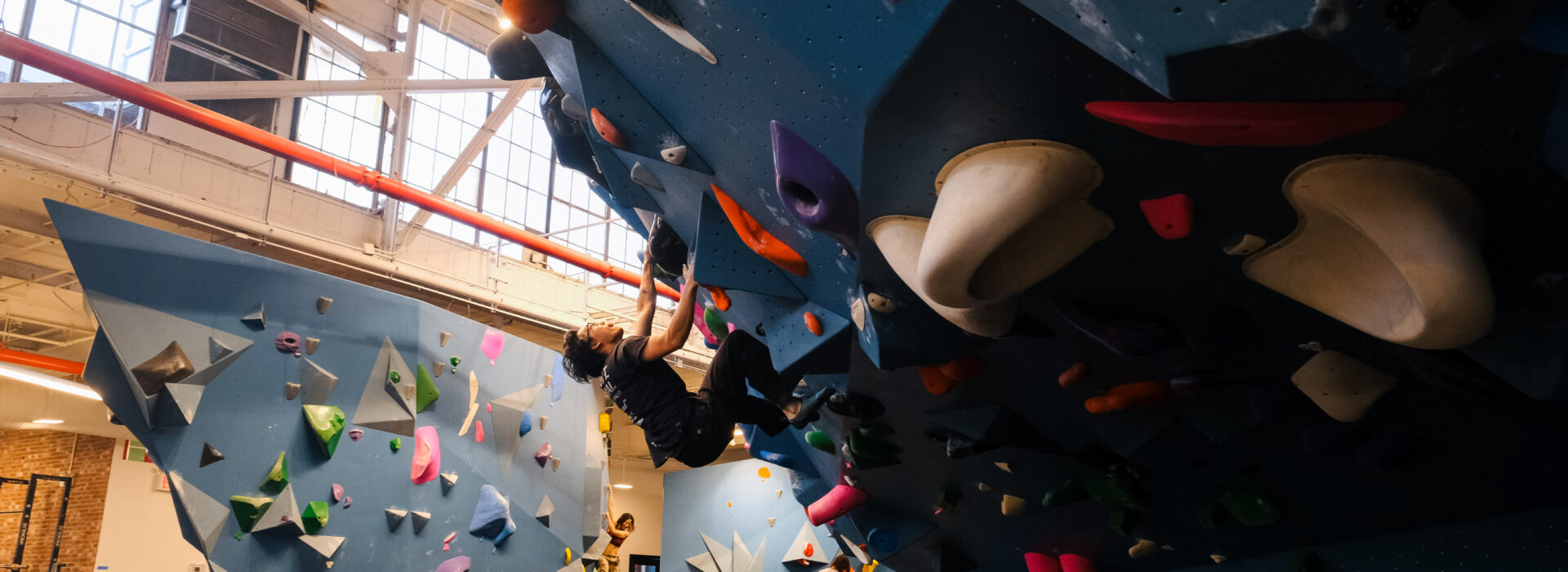 A person climbs a blue indoor bouldering wall covered with colorful holds at Brooklyn Climbing Yoga and Fitness, while others climb and observe in the spacious gym. Natural light streams through large windows above.