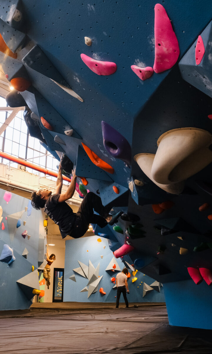 A person climbs a blue indoor bouldering wall covered with colorful holds at Brooklyn Climbing Yoga and Fitness, while others climb and observe in the spacious gym. Natural light streams through large windows above.