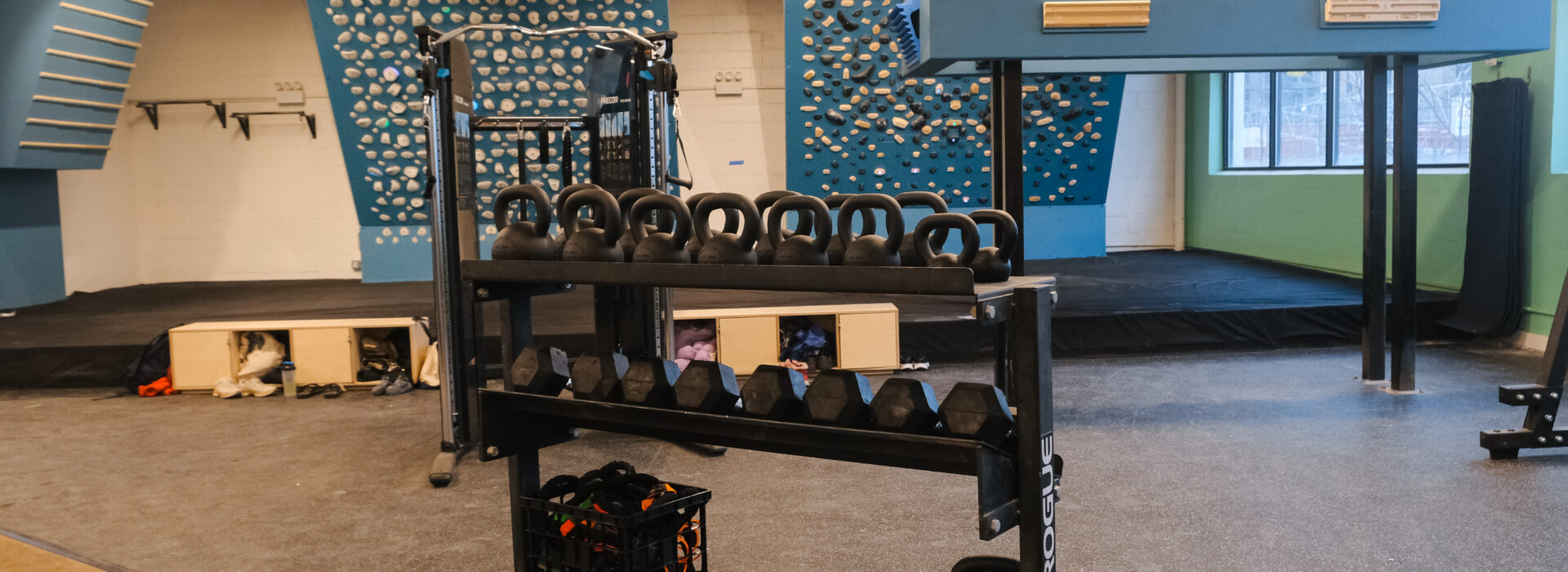A gym at Brooklyn Climbing Yoga and Fitness features a rack of kettlebells and free weights in the foreground, with indoor climbing walls, various holds, and training equipment visible in the background.