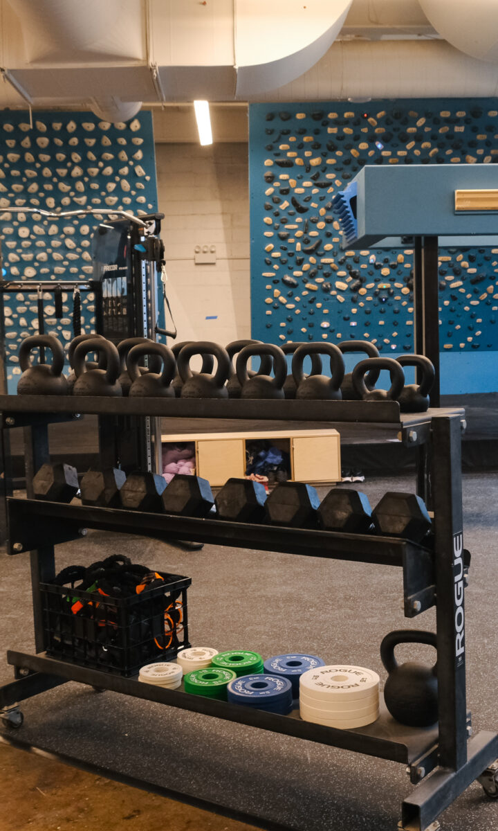 A gym at Brooklyn Climbing Yoga and Fitness features a rack of kettlebells and free weights in the foreground, with indoor climbing walls, various holds, and training equipment visible in the background.