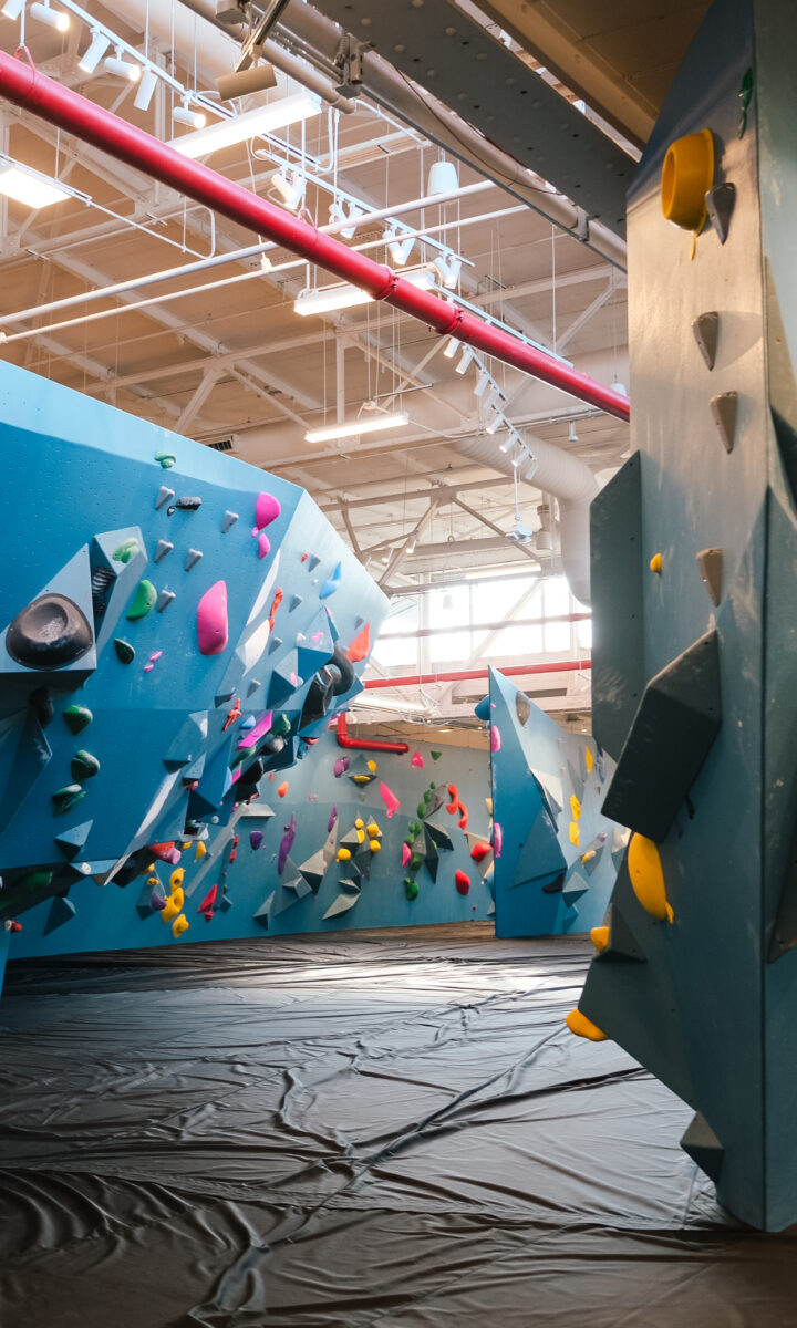 Indoor rock climbing gym with blue climbing walls, colorful holds, and black padded flooring beneath. Brooklyn Climbing Yoga and Fitness offers a well-lit space with overhead lights and multiple climbing routes of varying difficulty.