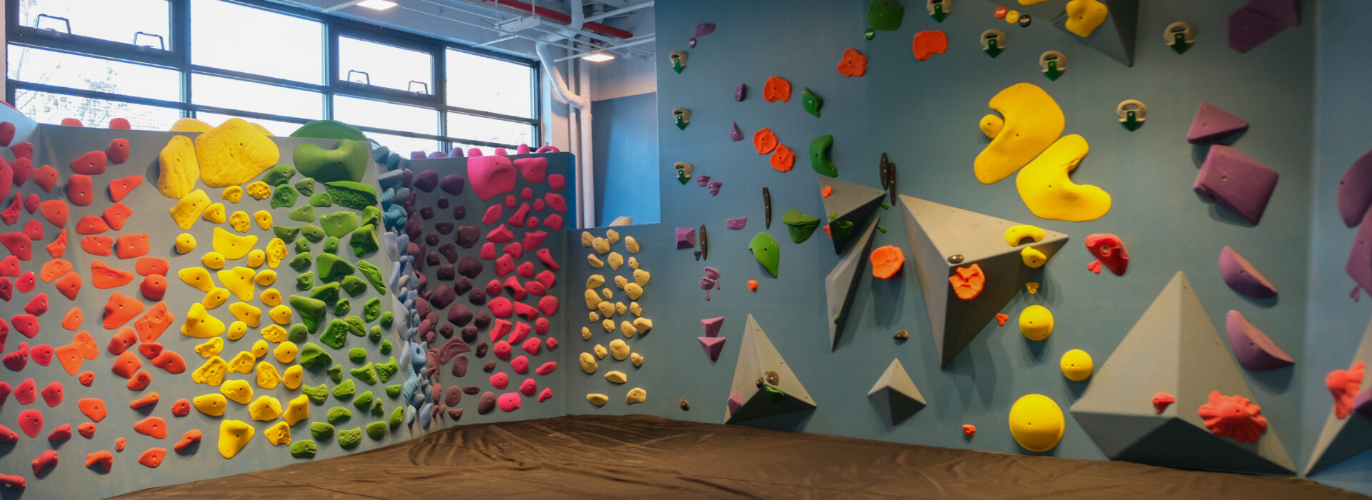 An indoor bouldering gym with colorful climbing holds arranged in patterns on blue walls, Brooklyn Climbing Yoga and Fitness also features a thick padded mat on the floor for safety.