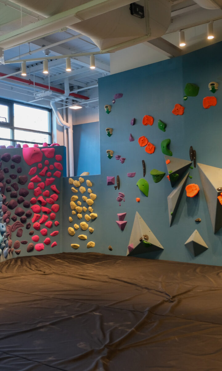 An indoor bouldering gym with colorful climbing holds arranged in patterns on blue walls, Brooklyn Climbing Yoga and Fitness also features a thick padded mat on the floor for safety.