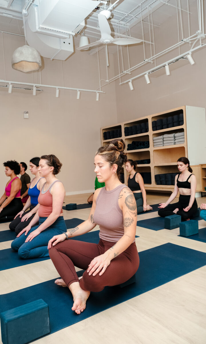 A group of people sit cross-legged on yoga mats at Brooklyn Climbing Yoga and Fitness, eyes closed and hands resting on their knees, practicing meditation. Yoga blocks are beside each mat, with shelves of rolled towels in the background.