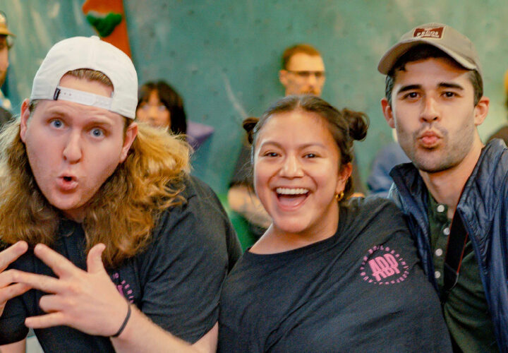 A group of people pose and smile for the camera in a lively indoor setting, with some making playful expressions and gestures. Others talk and laugh in the background, creating a cheerful, social atmosphere.