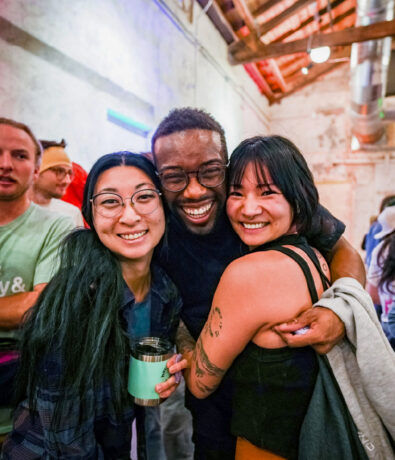 Three people stand close together indoors, smiling happily at the camera. The group is surrounded by others in the background, with exposed brick walls and beams visible above. One person in the center has their arms around the other two.
