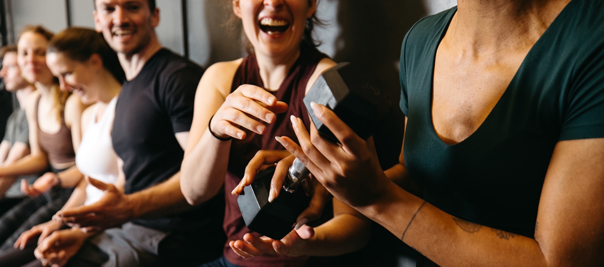 A group of people sit in a row, smiling and laughing together, as one person passes a dumbbell to another, creating a lively and friendly atmosphere often found in Fitness Classes.