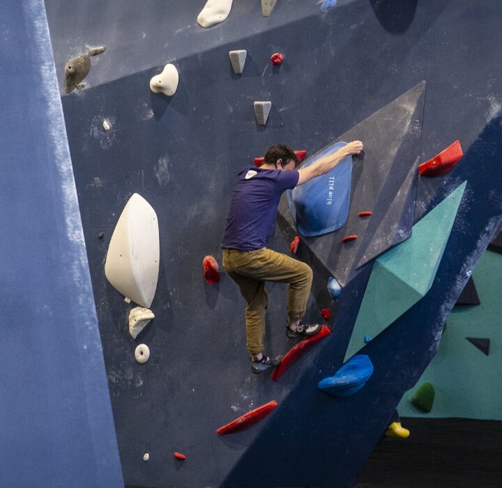 Three people are indoor rock climbing at Upper Walls in Fremont, tackling colorful, angled climbing walls with various shaped holds. Each climber follows a different route, using hands and feet to navigate the challenging surfaces.