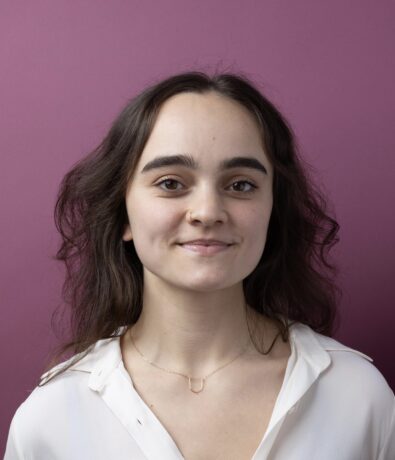 A young woman with long brown hair and a slight smile stands in front of a solid mauve background, wearing a white blouse and a delicate necklace—ready for her Brooklyn Climbing Yoga and Fitness class.