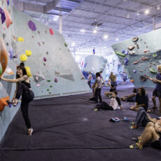 People bouldering and socializing inside a brightly-lit indoor climbing gym with large climbing walls, colorful holds, and a padded floor. Several climbers are on the wall while others watch or rest.