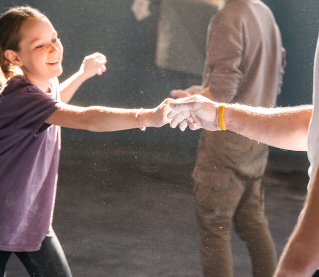 A smiling girl in a purple shirt reaches out to hold hands with an adult in a white shirt, both dusted with chalk, enjoying an After School Program in a brightly lit DC indoor setting, with another person blurred in the background.