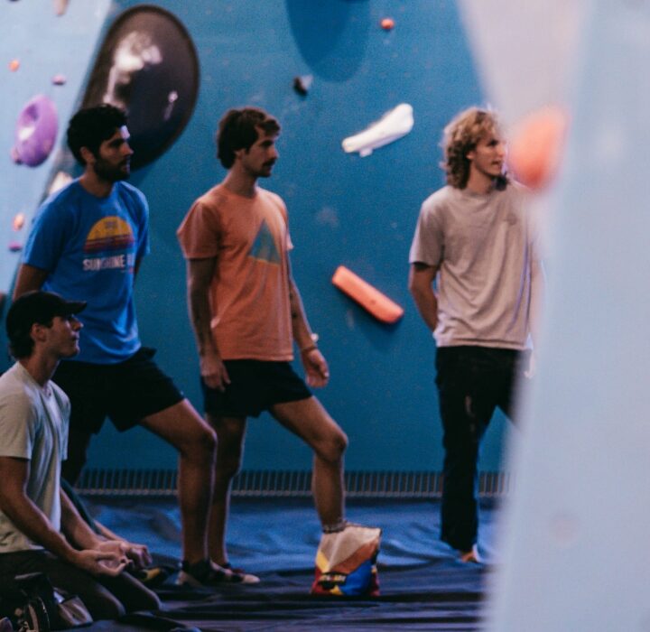A group of young people, some standing and some sitting, gather inside a climbing gym with colorful holds on blue walls. They appear to be watching or discussing something—perfect for those interested in group reservations.