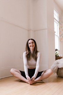 A woman sits on a wooden floor with her legs crossed and hands clasped in front of her, smiling. She is indoors in a bright room at a climbing gym in the University District, Seattle, with white walls and large windows.