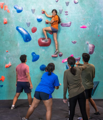 A woman climbs an indoor rock climbing gym wall as four people stand below, watching and encouraging her. The colorful climbing holds and supportive group create an engaging atmosphere.