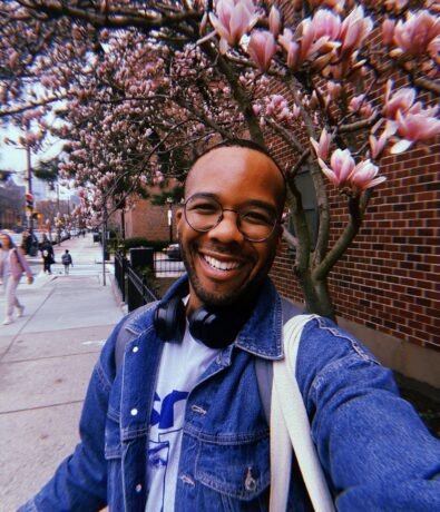 A smiling person wearing glasses, headphones, and a denim jacket takes a selfie outdoors near blooming pink magnolia trees and a brick building.