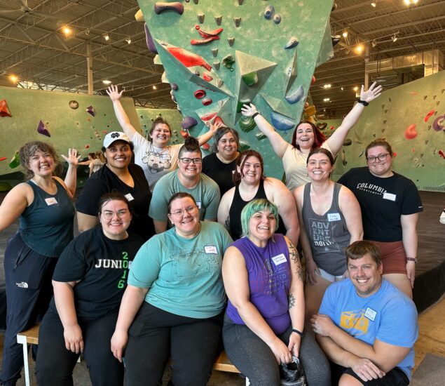 A group of smiling people from Community Clubs poses together in front of an indoor climbing wall in Minneapolis. Some sit on a bench, others stand behind with arms raised in excitement, dressed in casual athletic clothes and name tags.