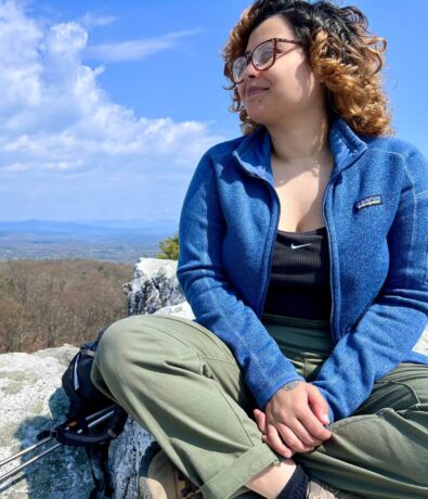 A woman with curly hair, glasses, and a blue jacket sits on a rocky ledge outdoors, looking into the distance. Behind her is a scenic view of trees and mountains—an inspiring moment for any Brooklyn Climbing Yoga and Fitness enthusiast.