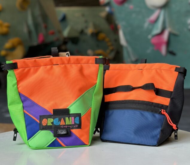 Two colorful chalk bags sit on a white surface, perfect for retail display. The left bag features orange, green, and purple geometric patterns with an Organic patch, while the right MBP bag is mostly orange with blue and black accents. A climbing wall blurs in the background.