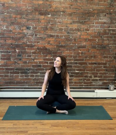 A woman in black workout clothes sits cross-legged on a green yoga mat in front of a brick wall, looking up and smiling. The room has hardwood floors and a white radiator along the wall.