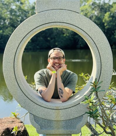 A smiling person with glasses and tattoos poses with their head resting on their hands inside a large stone circular sculpture by a pond, framed by lush trees—capturing peaceful moments after a day at the DC Rock Climbing Gym.