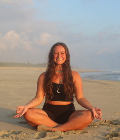 A woman sits cross-legged on a sandy Fremont beach, smiling and meditating with her hands resting on her knees. The ocean and blue sky with clouds create a serene background.