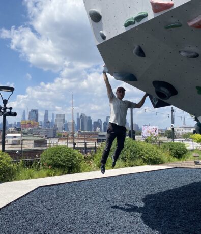 A person hangs by one arm from an outdoor climbing wall with colorful holds, with a city skyline and blue sky with clouds in the background.