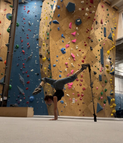 A person balances on their hands in a handstand split pose, with one foot on a support stick, in front of tall indoor climbing walls covered in colorful holds.