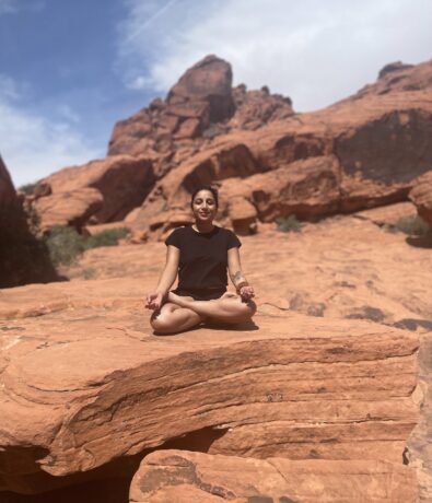 A person sits cross-legged in a meditative pose on a large red rock, reminiscent of the rugged formations near a climbing gym in Seattle’s University District, surrounded by red sandstone and a partly cloudy sky.