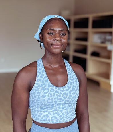 A woman wearing a blue patterned sports bra and matching headscarf stands in a bright Brooklyn Climbing Yoga and Fitness studio with wooden shelves, looking at the camera with a calm expression.