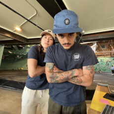 Two people stand indoors with arms crossed, both wearing blue shirts and hats. The person in front has tattoos on their arms and a mustache. Behind them is a climbing gym with colorful climbing holds and padded floors.