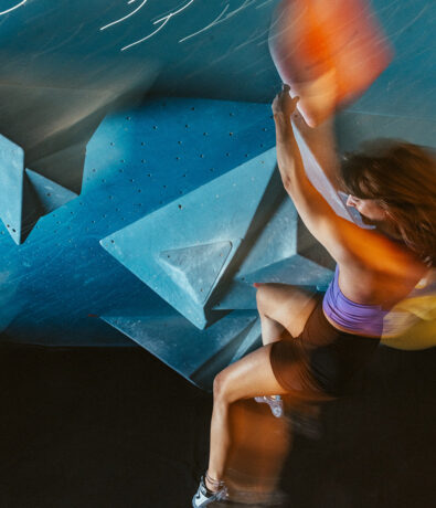 A woman in athletic wear climbs an indoor bouldering wall with large, angular blue holds. Her movements are blurred, suggesting rapid action and intensity.