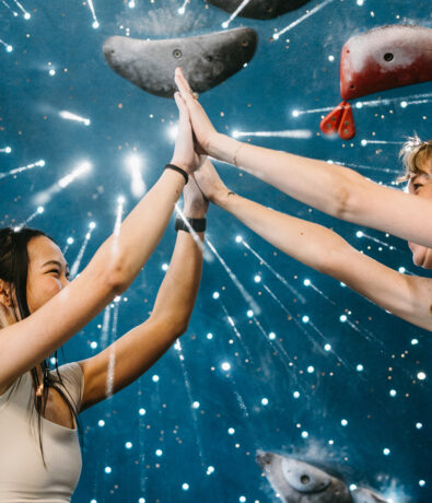 Two people smiling and high-fiving each other in front of a colorful indoor climbing wall with handholds and a starburst design in the background.