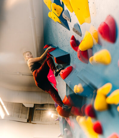 A person in a tank top and red pants climbs an indoor bouldering wall at a climbing gym in the University District, Seattle, gripping a blue hold and looking upward amid colorful holds in the well-lit space.